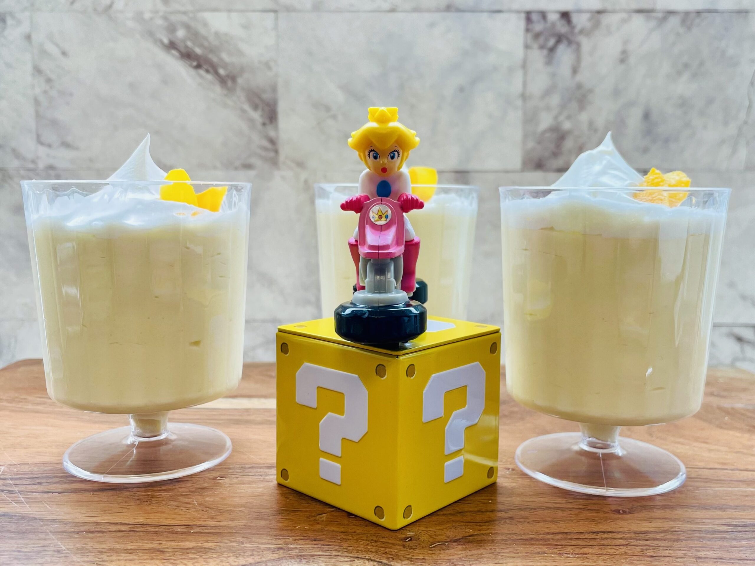 La imagen muestra tres copas con mousse de durazno con crema para batir y durazno en la parte superior. En medio hay una cubierta de superpoderes de Mario Broa con una figura de la princesa Peach en una moto.