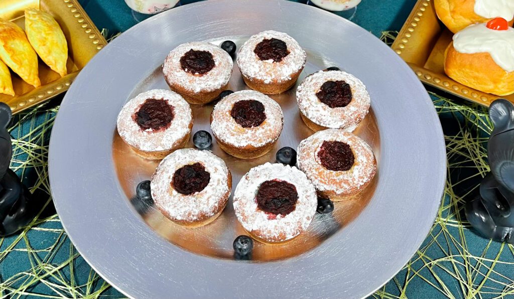 La imagen muestra un plato color plateado con pastelitos de Valiente, hechos con muffins rellenos de crema con Moras azules, decorados con mermelada de Moras y espolvoreados con azúcar glass.