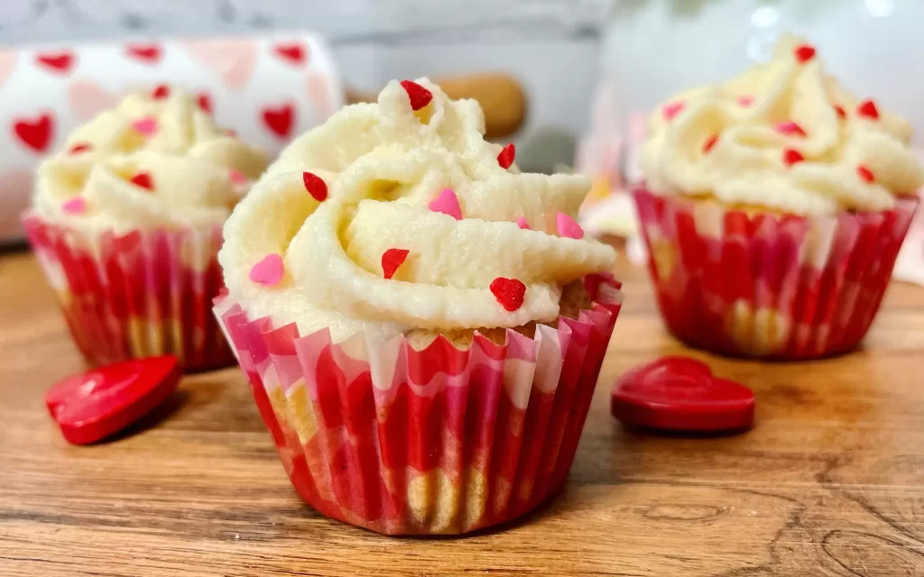 text=Tres mini cupcakes de fresa con glaseado de queso crema y chispas rojas y rosas en forma de corazón, con capacillos rojos, sobre una tabla de madera enfrente de un rodillo blanco con corazones rosas y rojos
