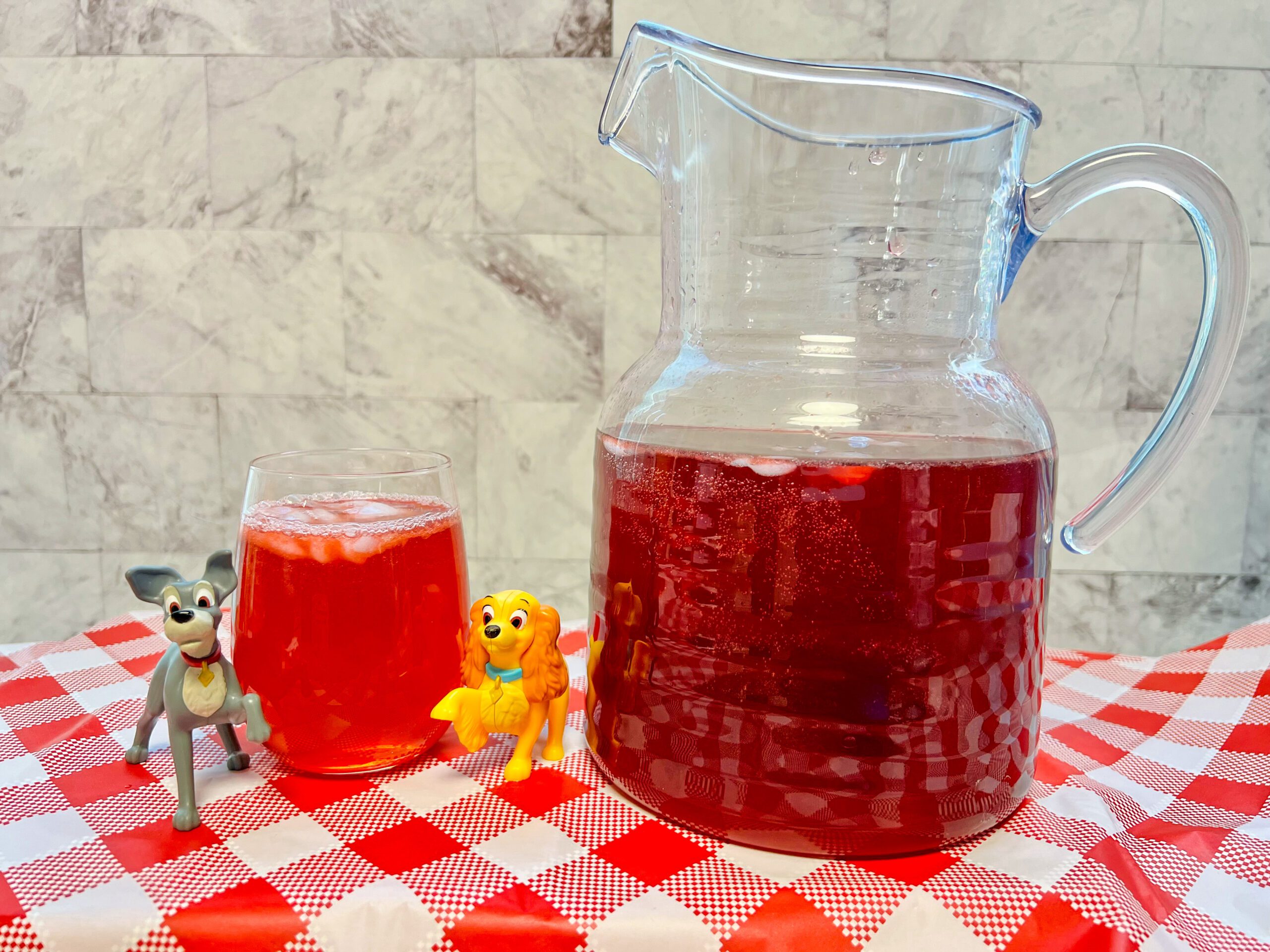 La imagen muestra un vaso de bebida de uva-arándano inspirada en la Dama y el Vagabundo, junto a una jarra.