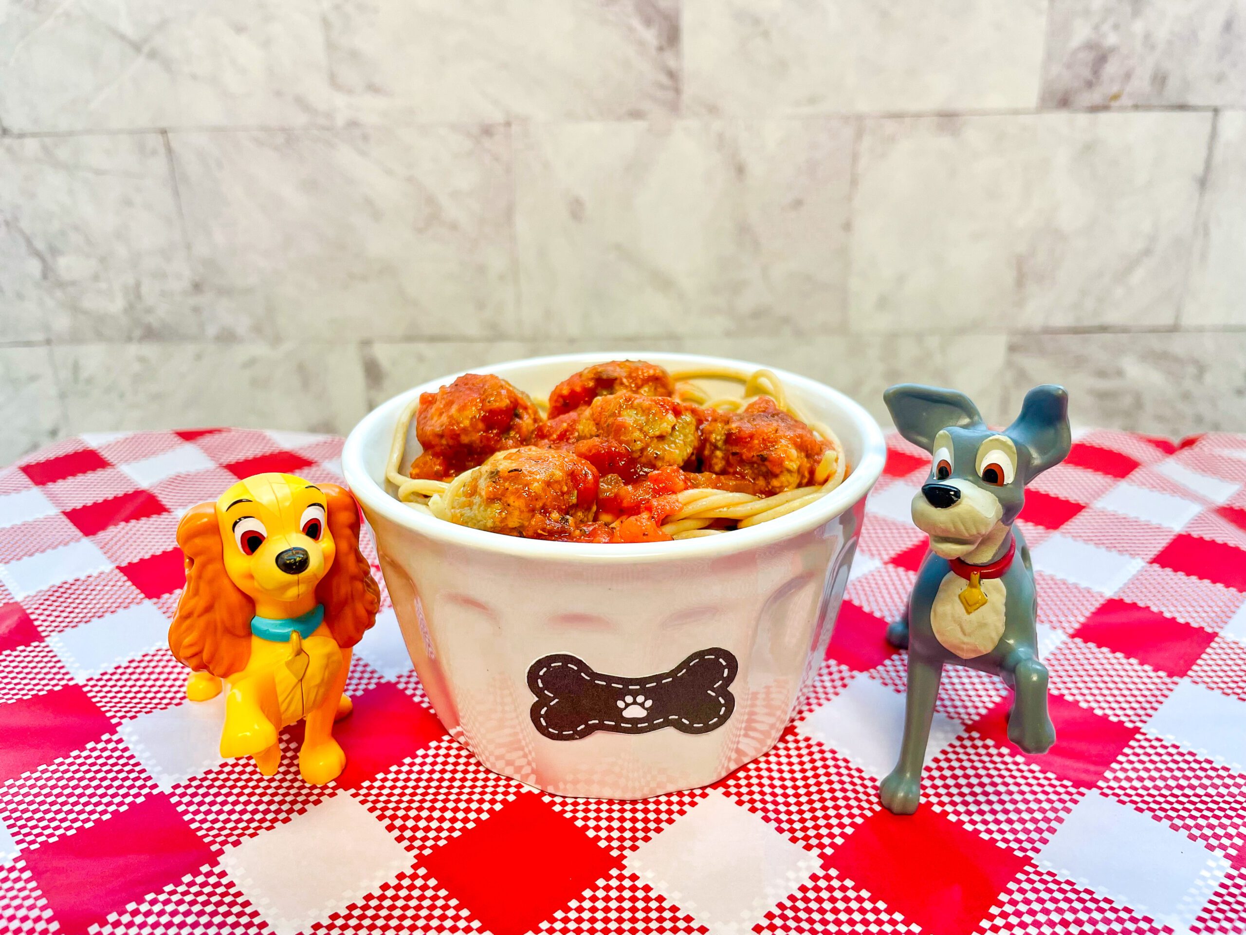 La imagen muestra un ramekin con una figura de hueso con espagueti con albóndigas, en medio de unas figuras de juguete de la Dama y el Vagabundo, sobre un mantel de cuadros rojos y blancos.