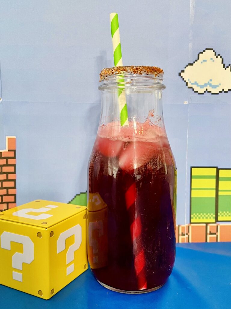 La imagen muestra una bebida de flor de fuego inspirada en Mario Bros, hecha con agua de jamaica y limón, servida en una botella de vidrio escarchada con chile piquín, con un background del mundo de Mario Bros.