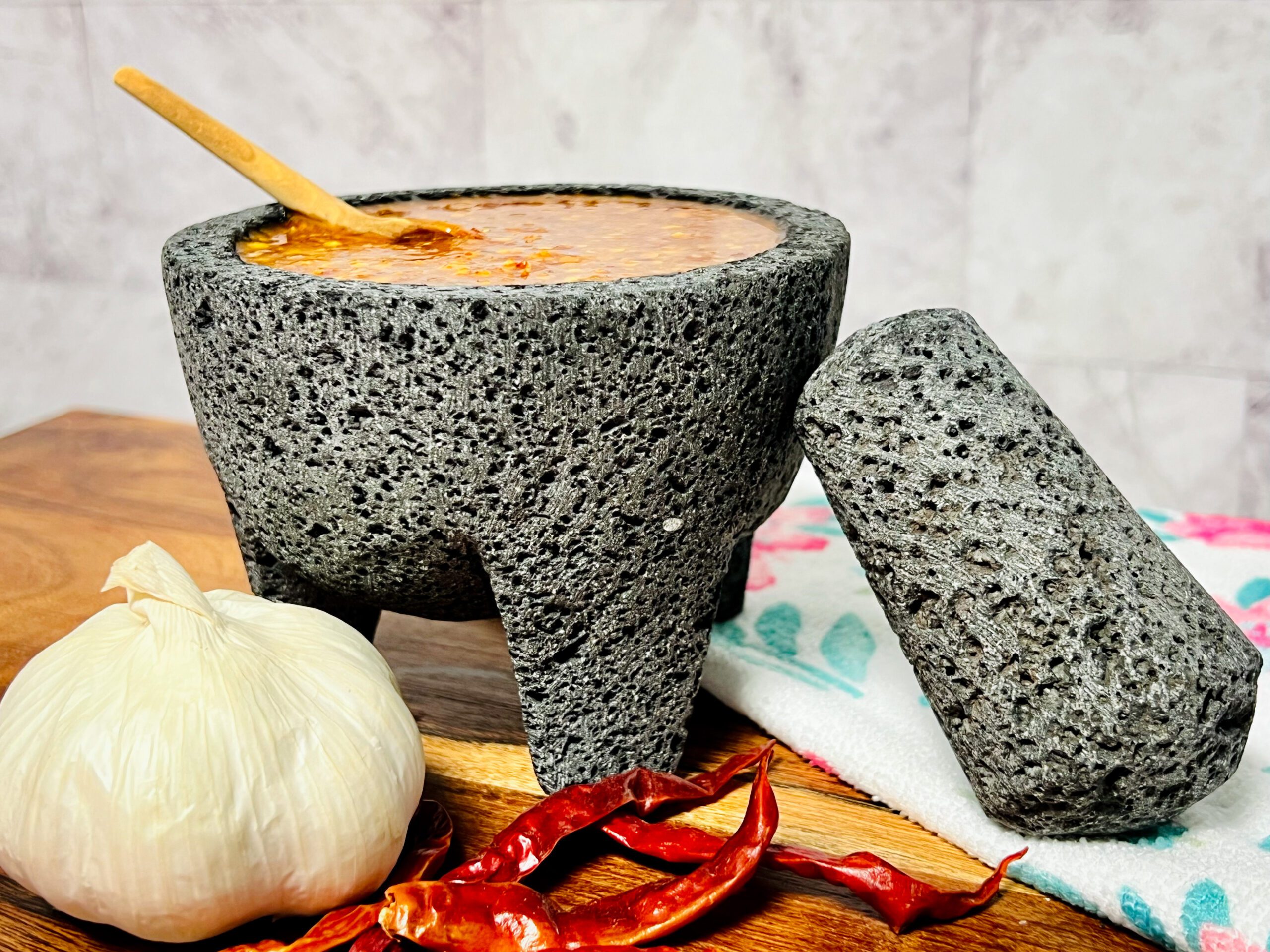 La imagen muestra un molcajetero con salsa de chiles de árbol y ajonjolí, con una cuchara de madera pequeña adentro, la piedra del molcajetero esta recargada en él y en frente hay unos chiles de árbol y un ajo.