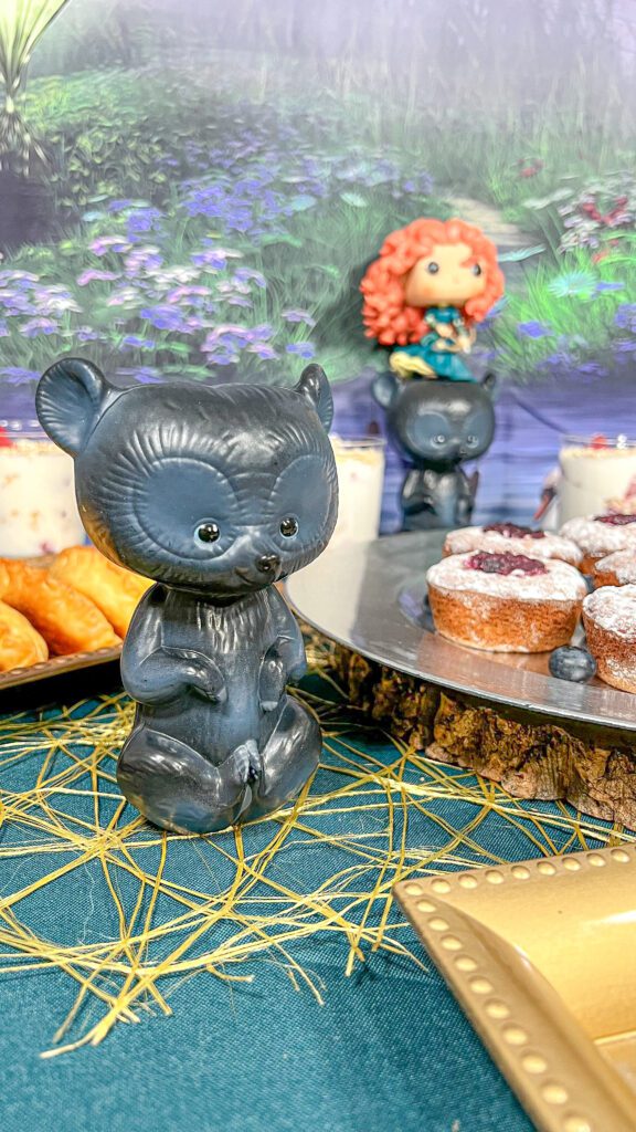 La imagen muestra una mesa con decoración y comida inspiradas en la película Valiente (Brave). La mes tiene un mantel verde con un camino de mesa dorado. Un plato sobre una base de madera con muffins que simulan los “pastelitos del hechizo” juntos 
