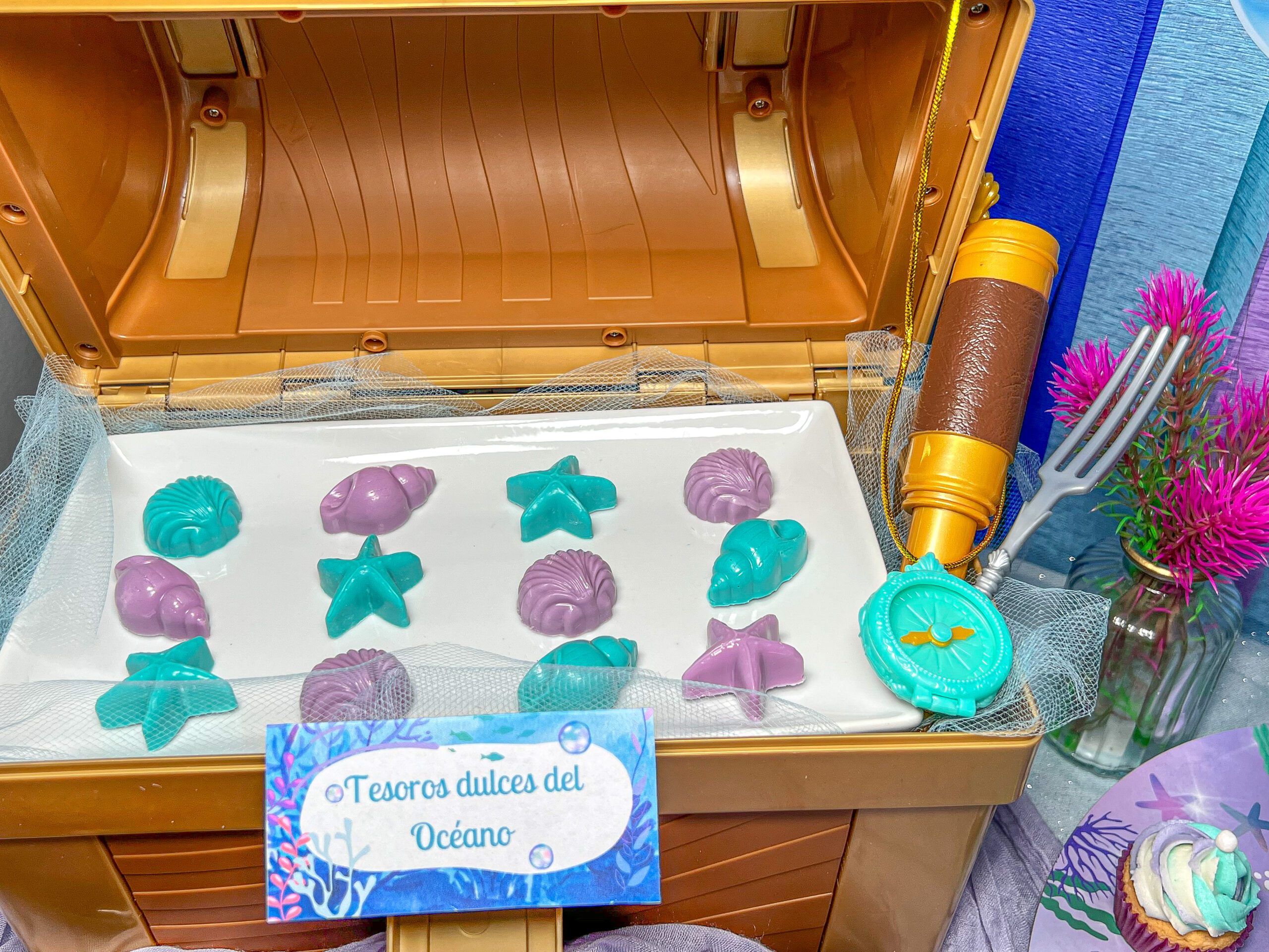Imagen de tesoros dulces del océano firmados con conchas marinas, caracoles y estrellas de mar de color turquesa y morado, en un plato rectangular dentro de un cofre de tesoros.