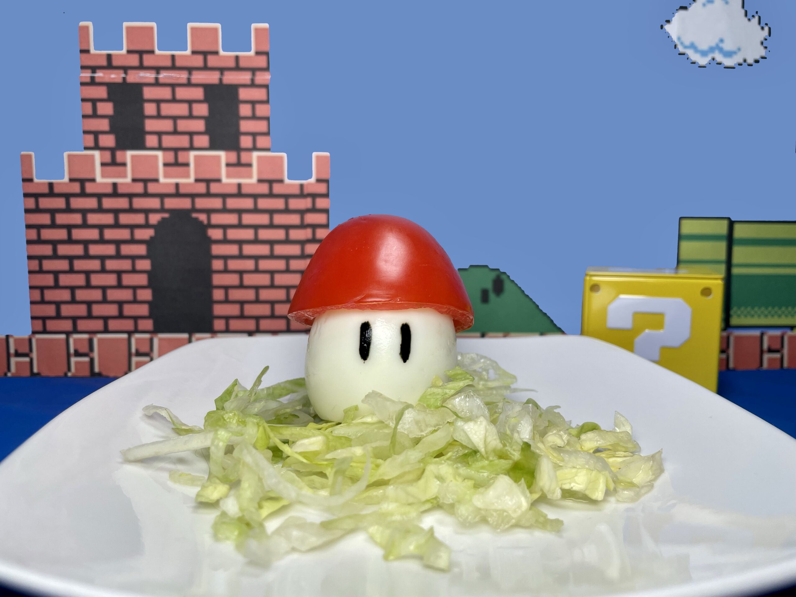 La imagen muestra un honguito de huevo y jitomate sobre una cama de lechuga con un fondo del castillo de Mario Bros.