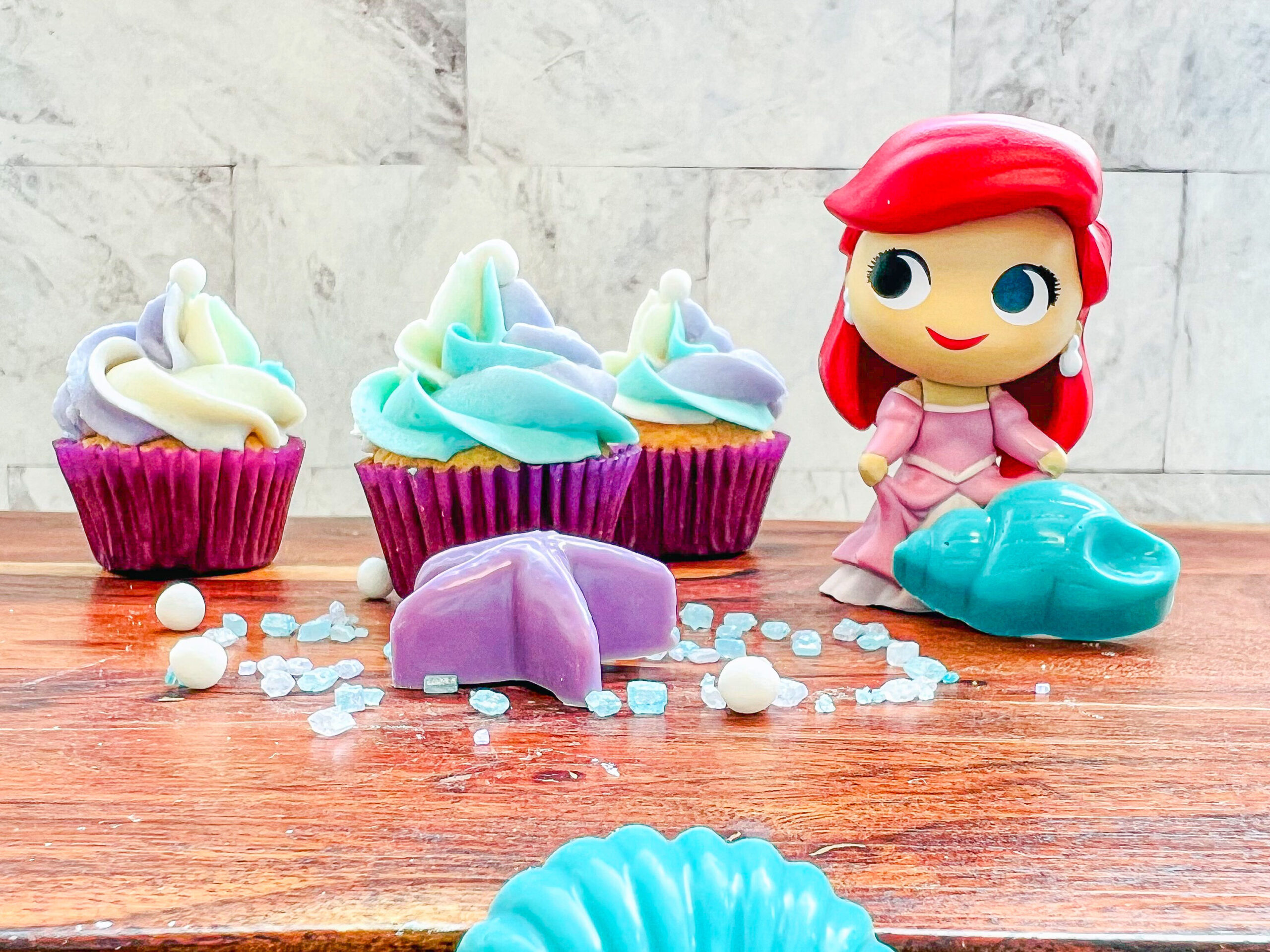 Imagen de 3 mini cupcakes con capacillos morados y glaseado de 3 colores: lila, blanco y turquesa, con una pela comestible en la parte superior. Se encuentran junto a una mini figura de Ariel y un caracol y una estrella de mar de caramelo. 