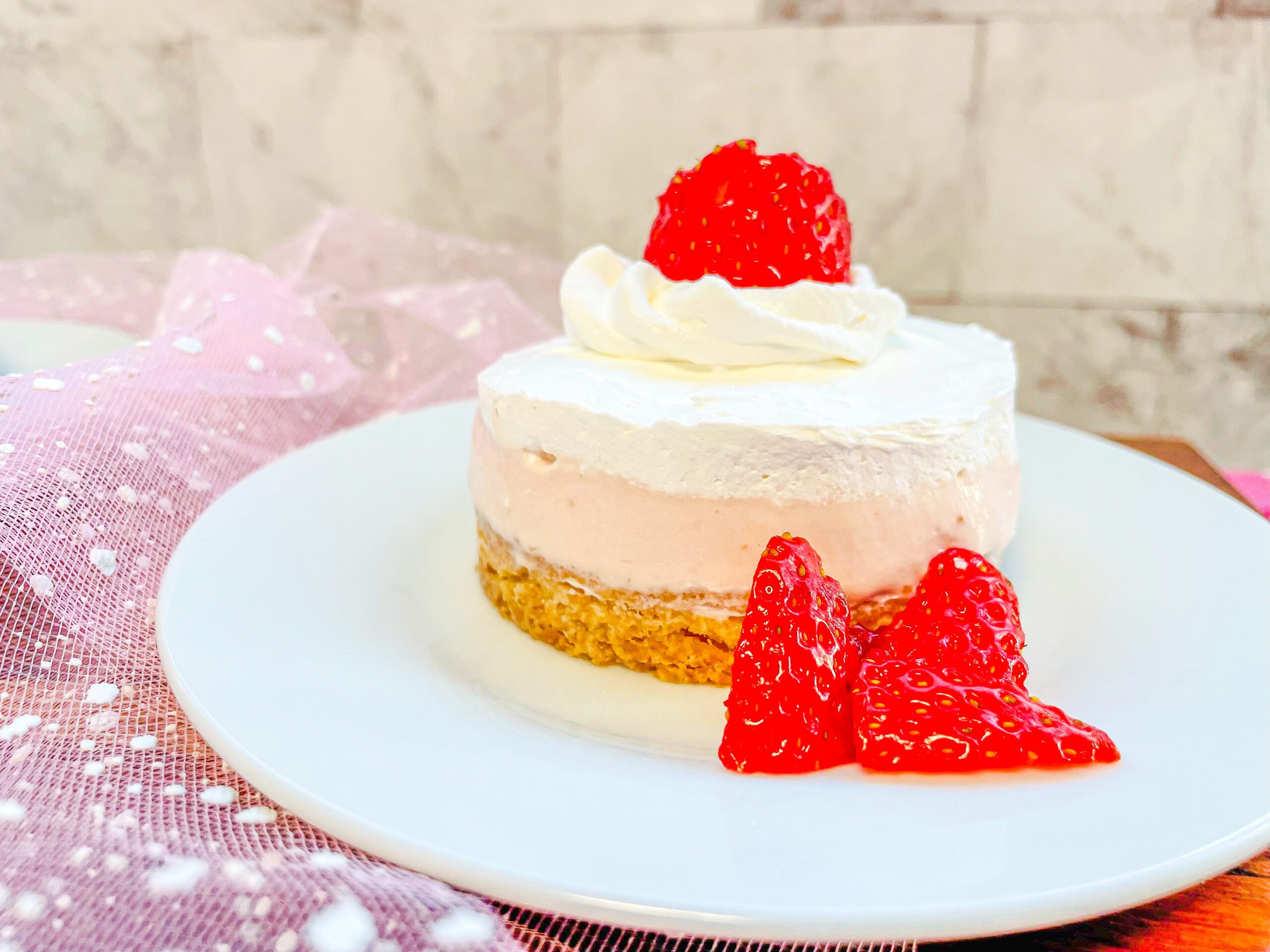 La imagen muestra un mini Mousse  de fresa con base de galleta de harina y una fresa libre crema batida en la parte superior. El Mousse está sobre un plato blanco redondo y 3 rebanadas de fresa al pie del Mousse para decorar. 