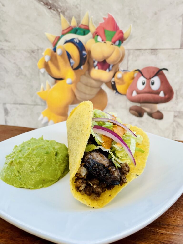 Taco de carne con champiñones acompañado con guacamolem en el fondo una imagen de Bowser con un hongo