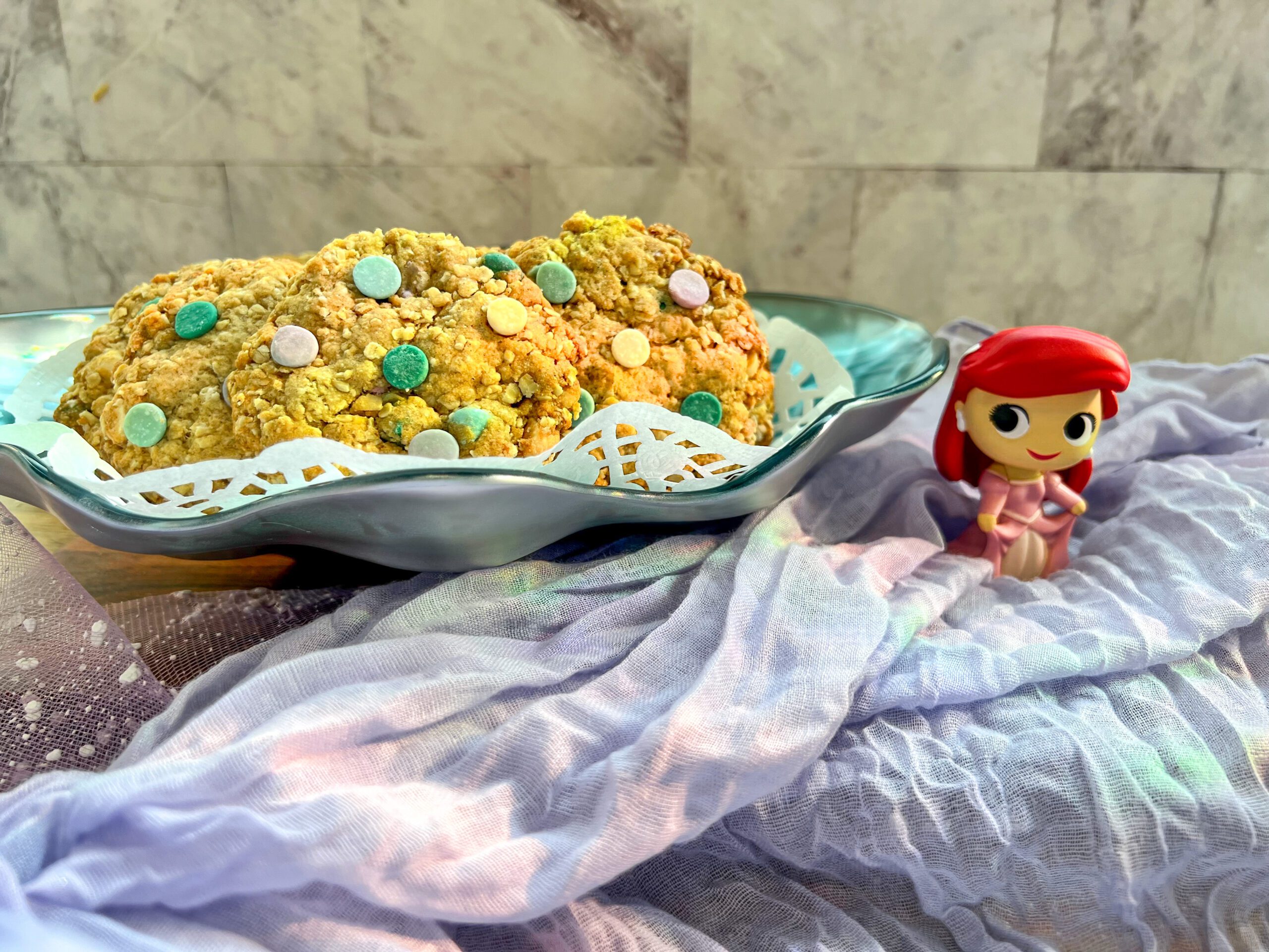Imagen de unas galletas de avena con chispas de colores lila, turquesa, verde y blanco en un recipiente con forma de concha marina. Sobre una superficie de madera con un camino de mesa lila y una figura de La Sirenita.