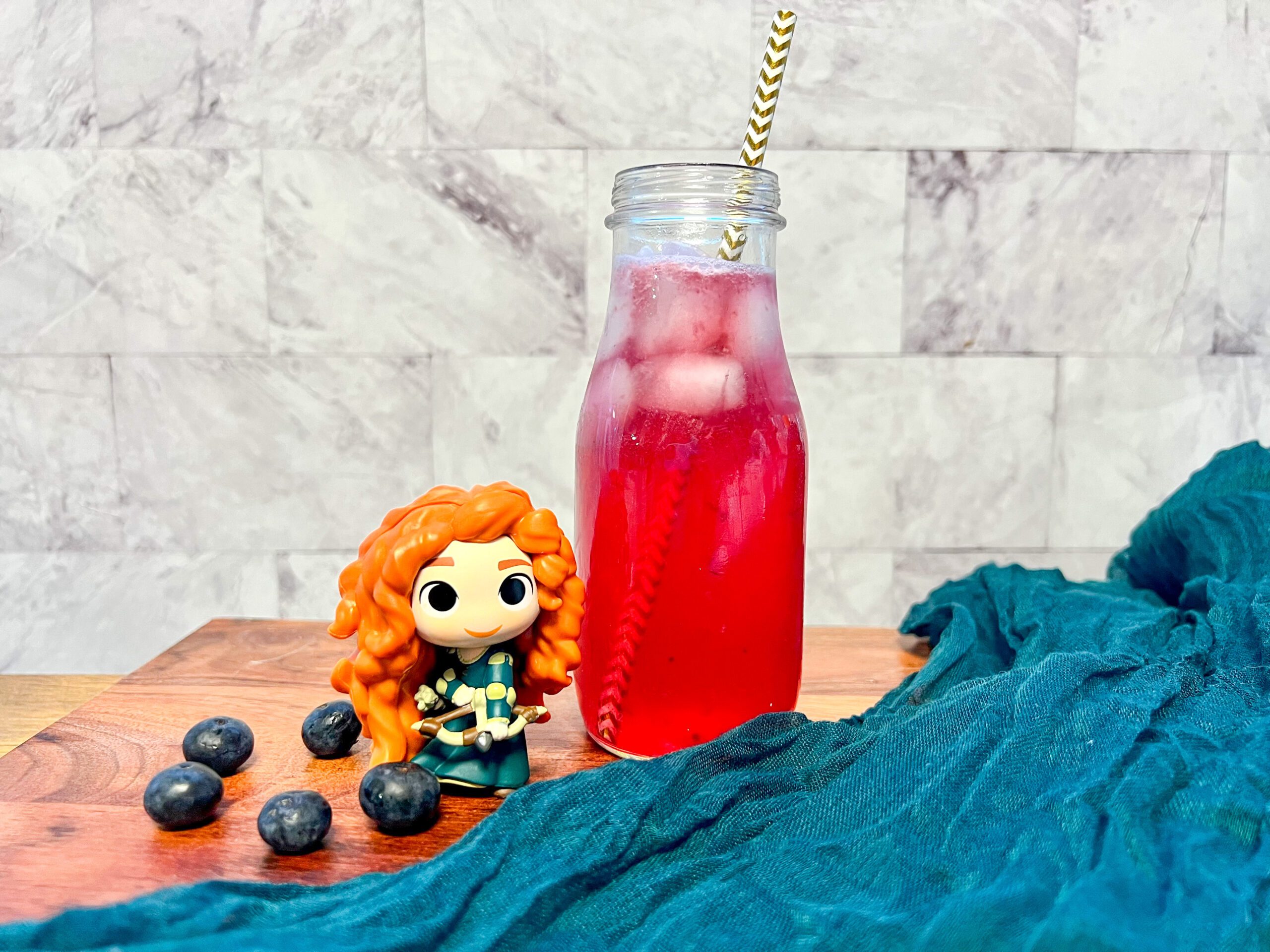 La imagen muestra una limonada de moras azules en una botella de vidrio con un popote con líneas blancas y doradas junto a una figura de funko de Merida y unas moras azules sobre una tabla de madera y un camino de mesa color azul verde.