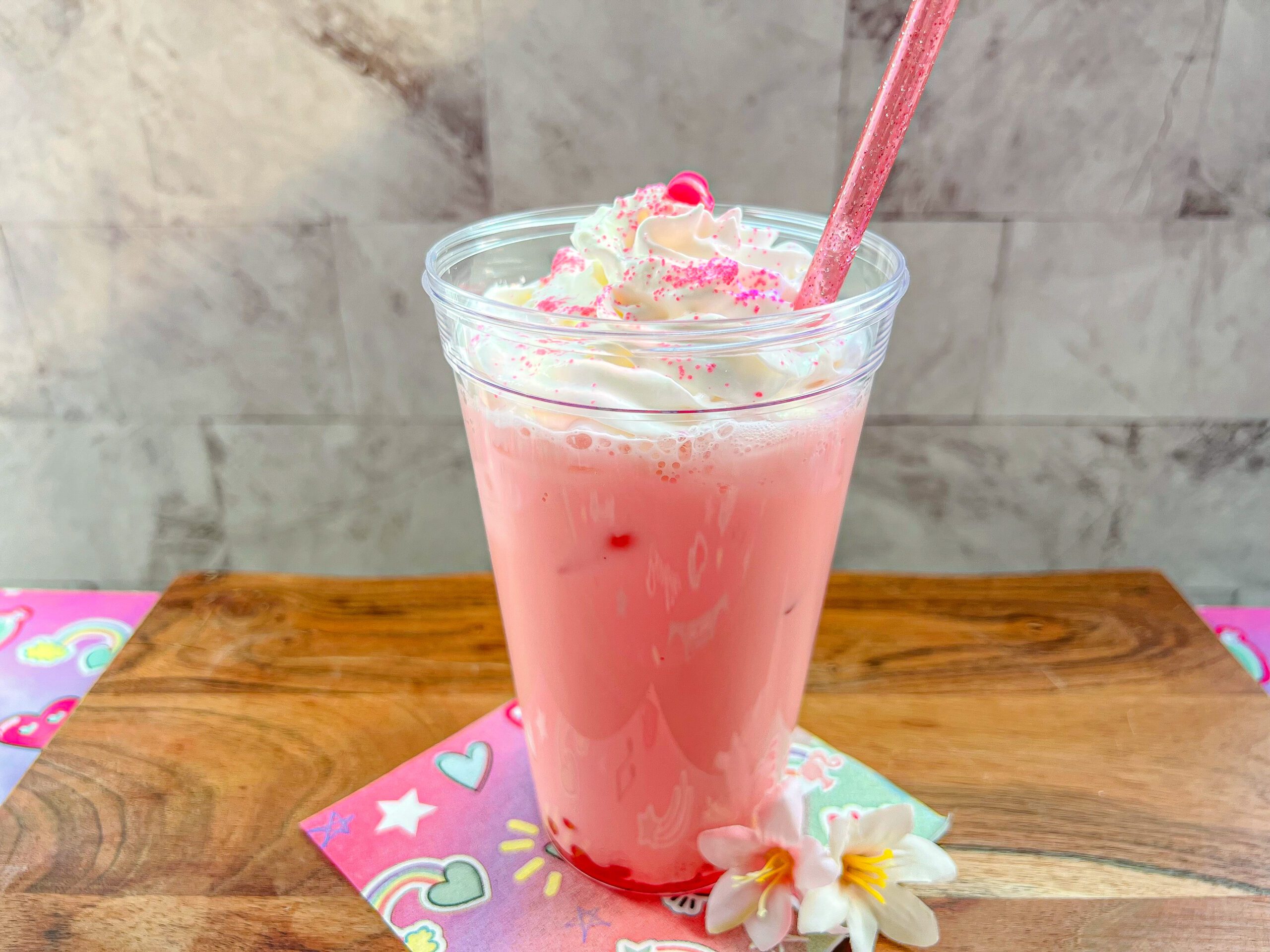 La imagen muestra un vaso con bebida rosa de Barbie, popping de fresa en el fondo, crema batida y azúcar rosa espolvoreada y un popote rosa con brillantina.