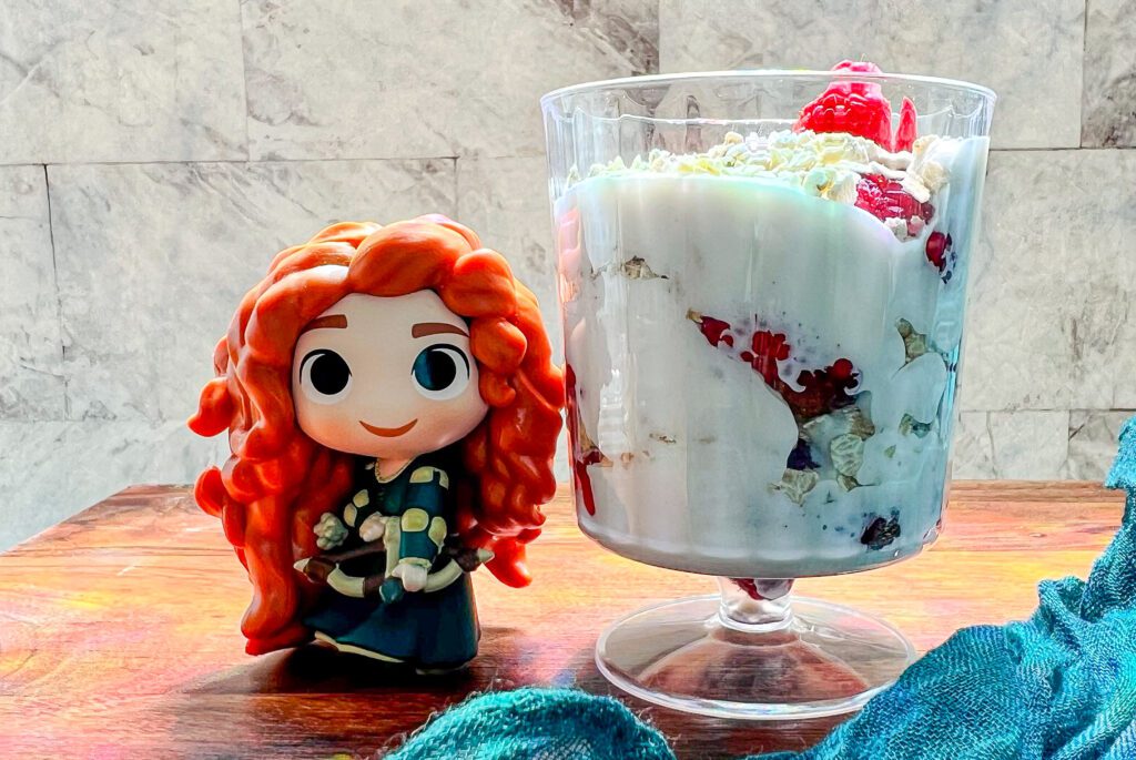 La imagen muestra una copa de postre de frambuesas con copos de avena inspirado en l película de Dante Vliente, junto a una figura de Merida.