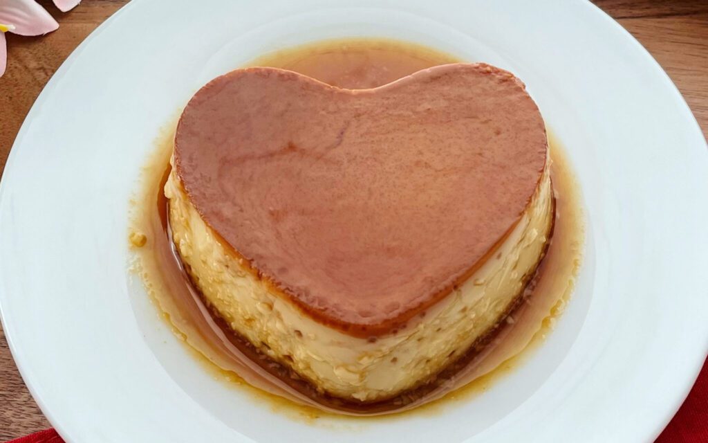 La imagen muestra un flan casero emn forma de corazón, bañado con caramelo, sobre un plato blanco, en una mesa de madera.