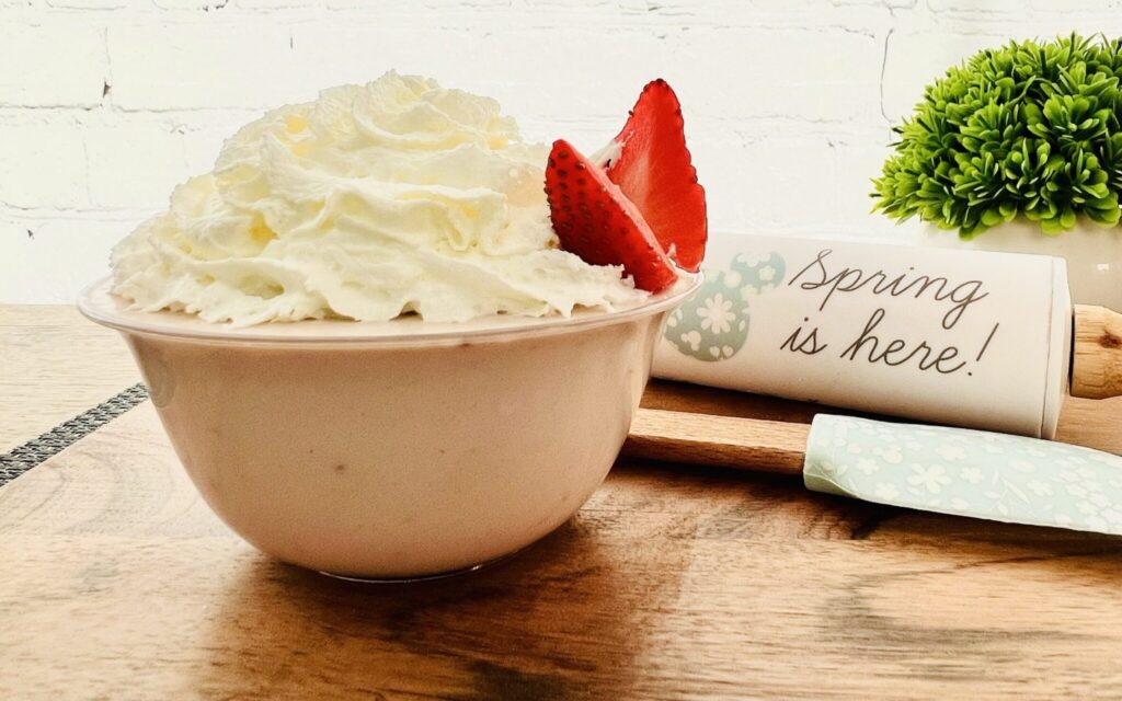 Imagen de mousse de fresa en un recipiente transparente, decorado con crema batida y 2 rebanadas de fresa que simulan las alas de una mariposa, el postre esta sobre una tabla de madera y puede verse un rodillo que dice "la,primavera está aqu"