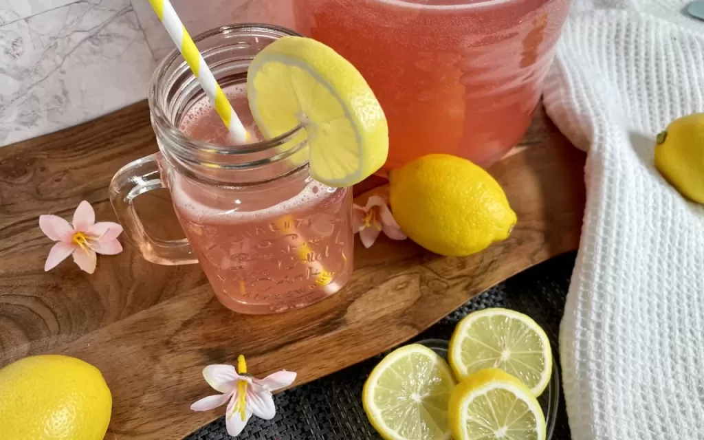Jarra de limonada rosa junto a un vaso con limonada rosa con un popote de papel blanco y amarillo decorado con una rodaja de limón amarillo, sobre una tabla de madera, con tres rebanadas de limón amarillo en frente.