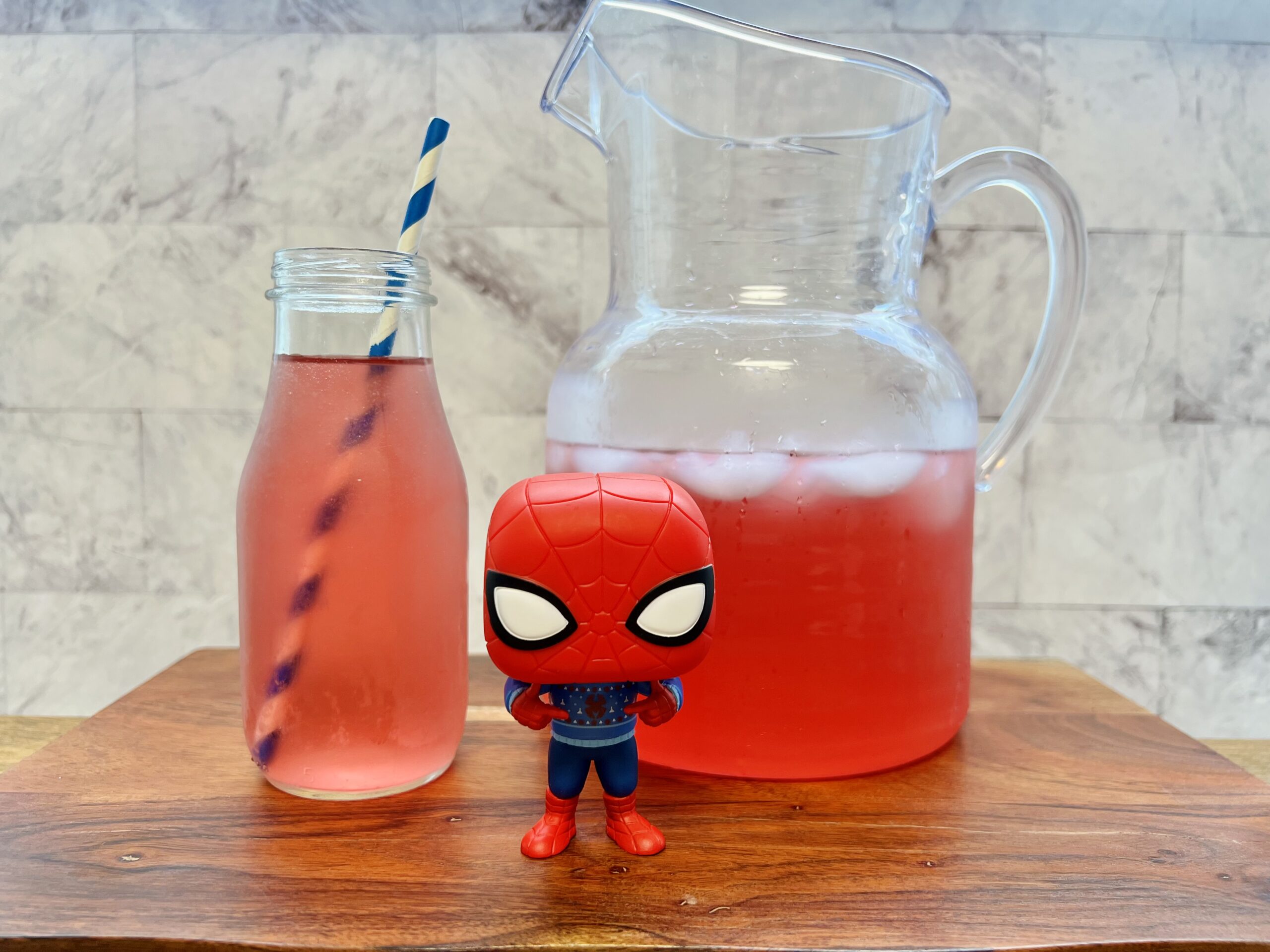 La imagen muestra una jarra y un vaso con ponche de frutas arácnido con un popote azul, junto a una figura de Spiderman. 