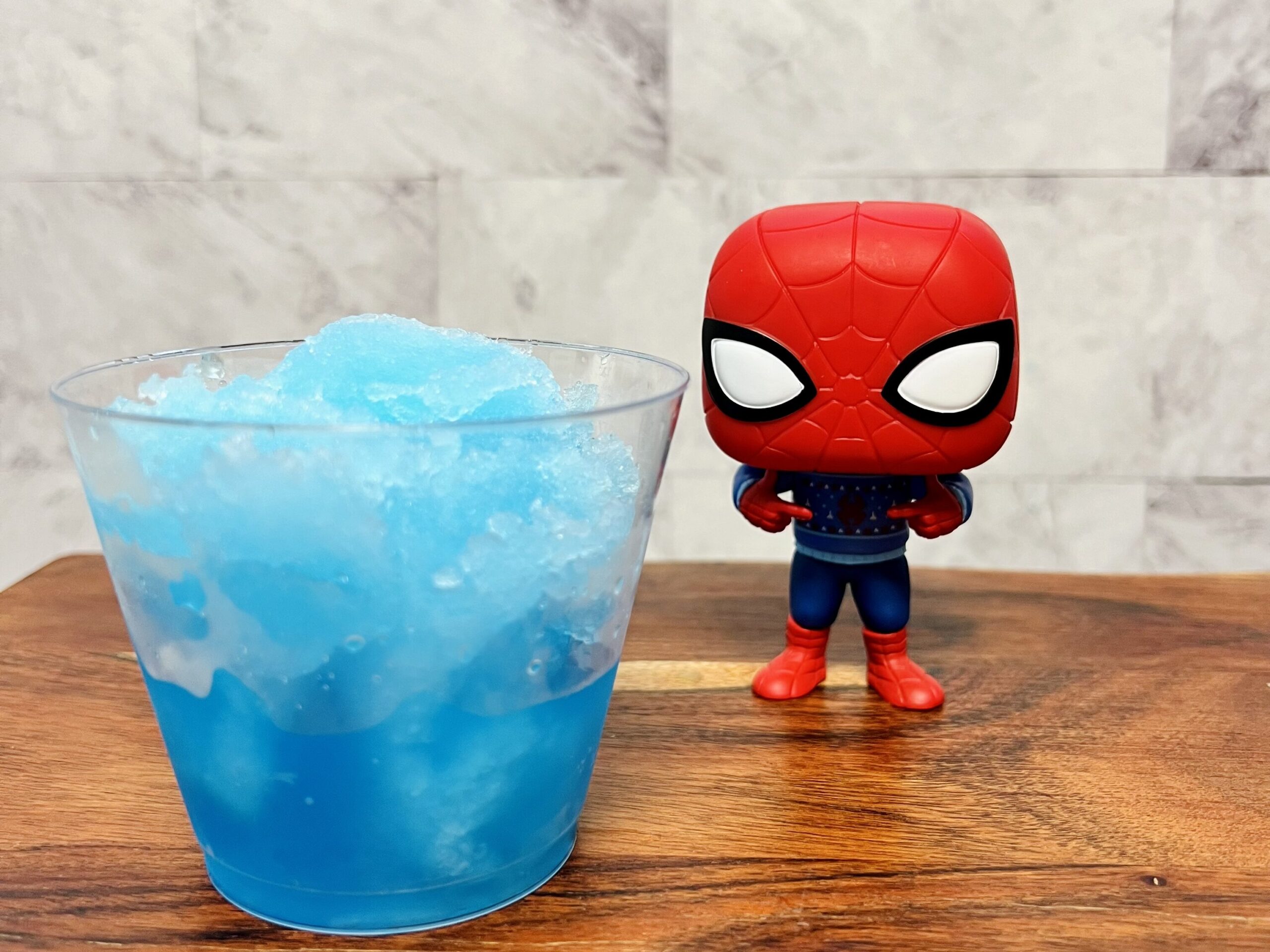 Imagen de un Spider-slushie (granizado azul) en un vaso transparente, junto a una figura de Funko de Spiderman.