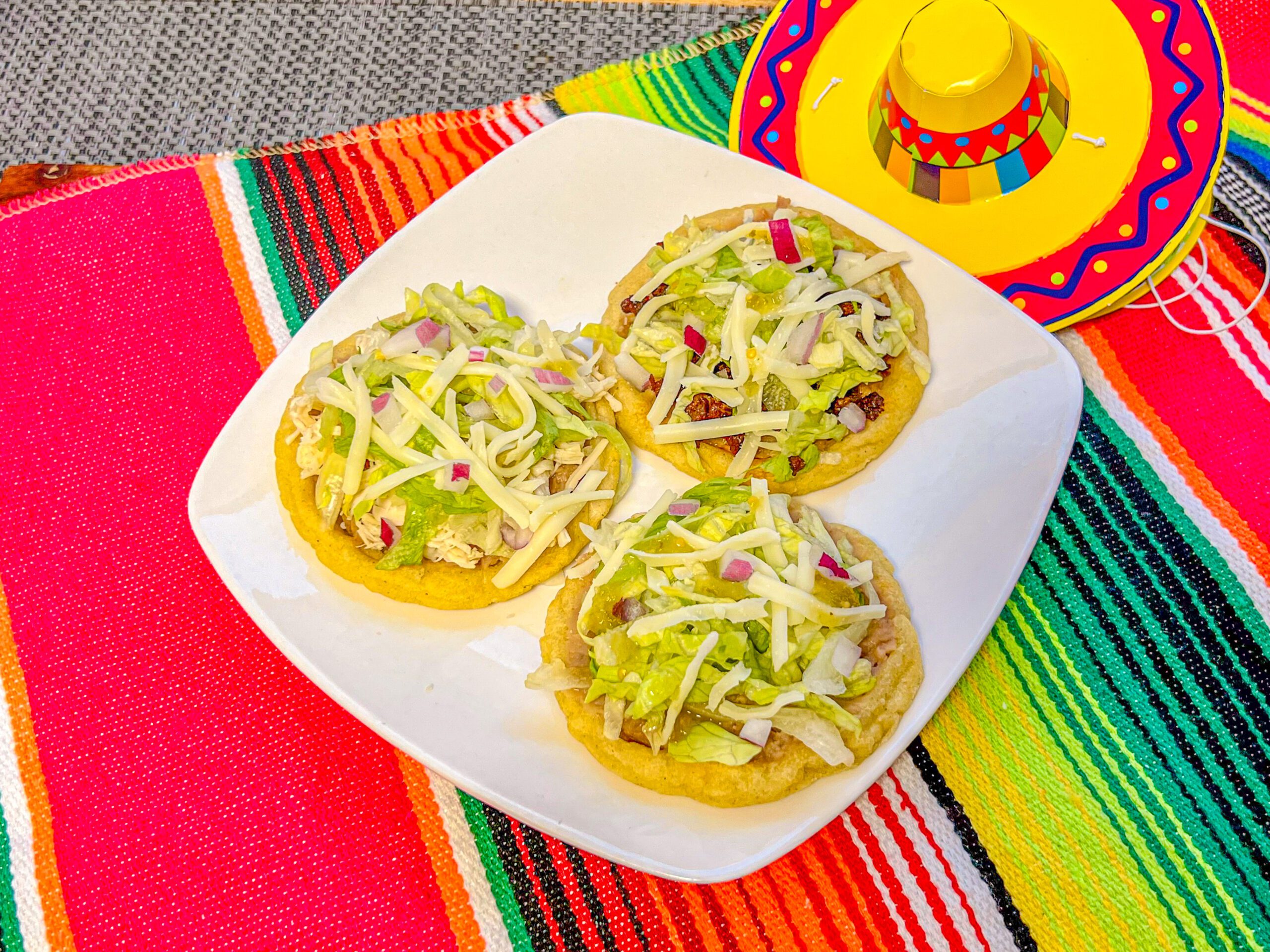 La imagen muestra un plato con 3 sopes de pollo y chorizo con lechuga, queso y cebolla morada, sobre un camino de mesa de sarape.