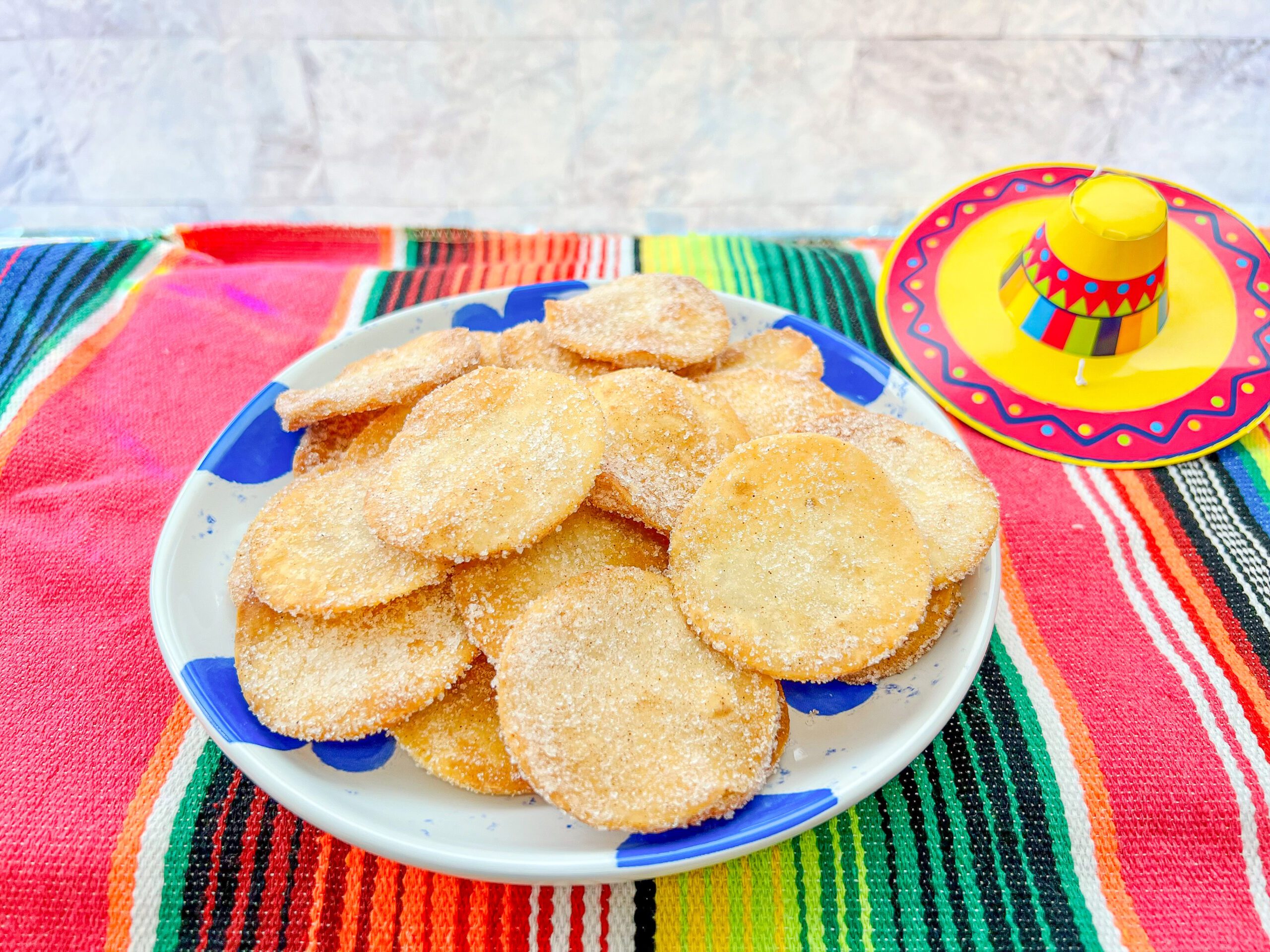 La imagen muestra un plato redondo con motivos azul rey con mini buñuelos hechos con tortillas de harina y cubiertos con azúcar y canela sobre un camino de mesa de sarape.