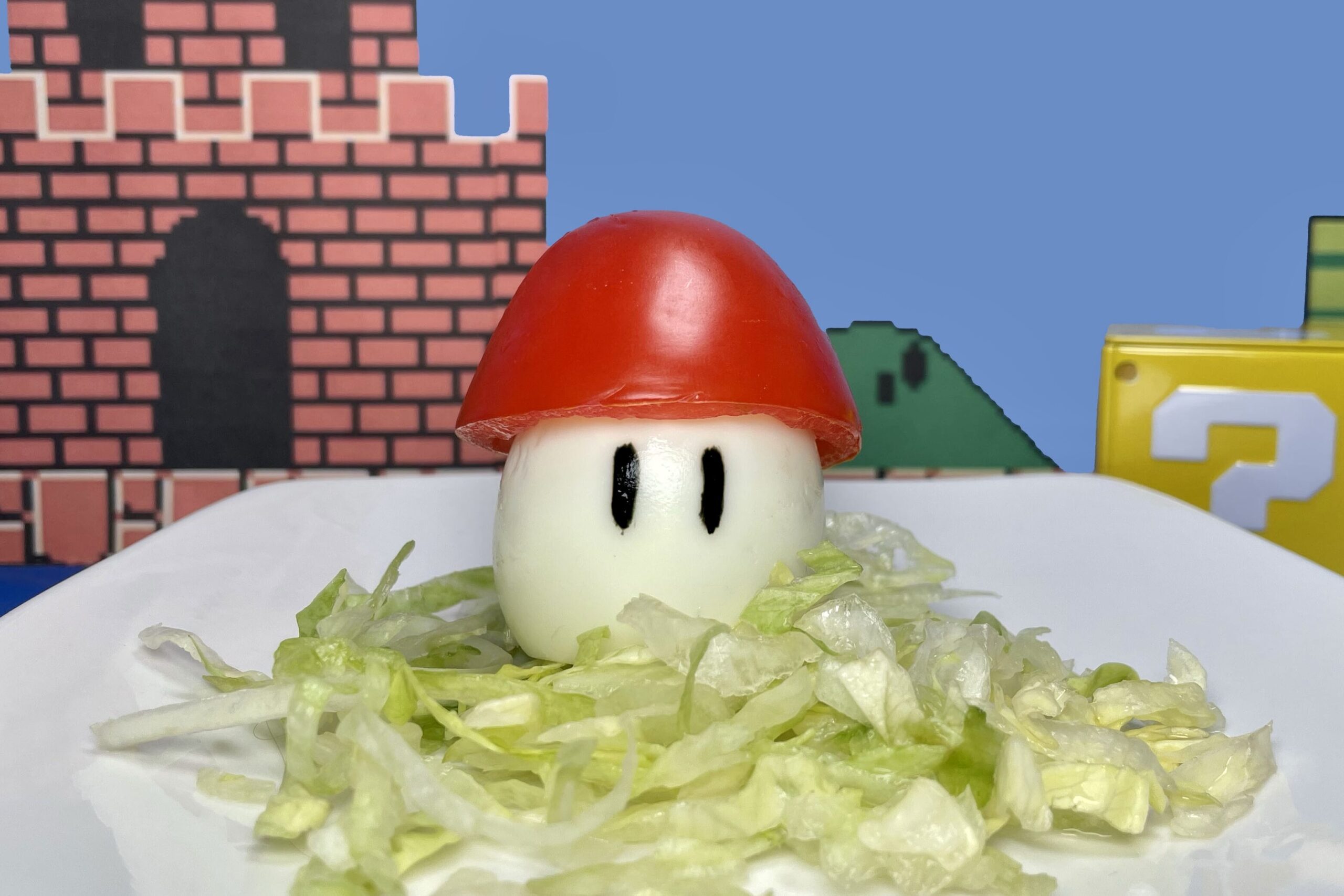 Honguito de Mario Bros hecho de huevo con sombrero de jitomate acompañado de lechuga, en el fondo se muestra el castillo y un cubo de pregunta