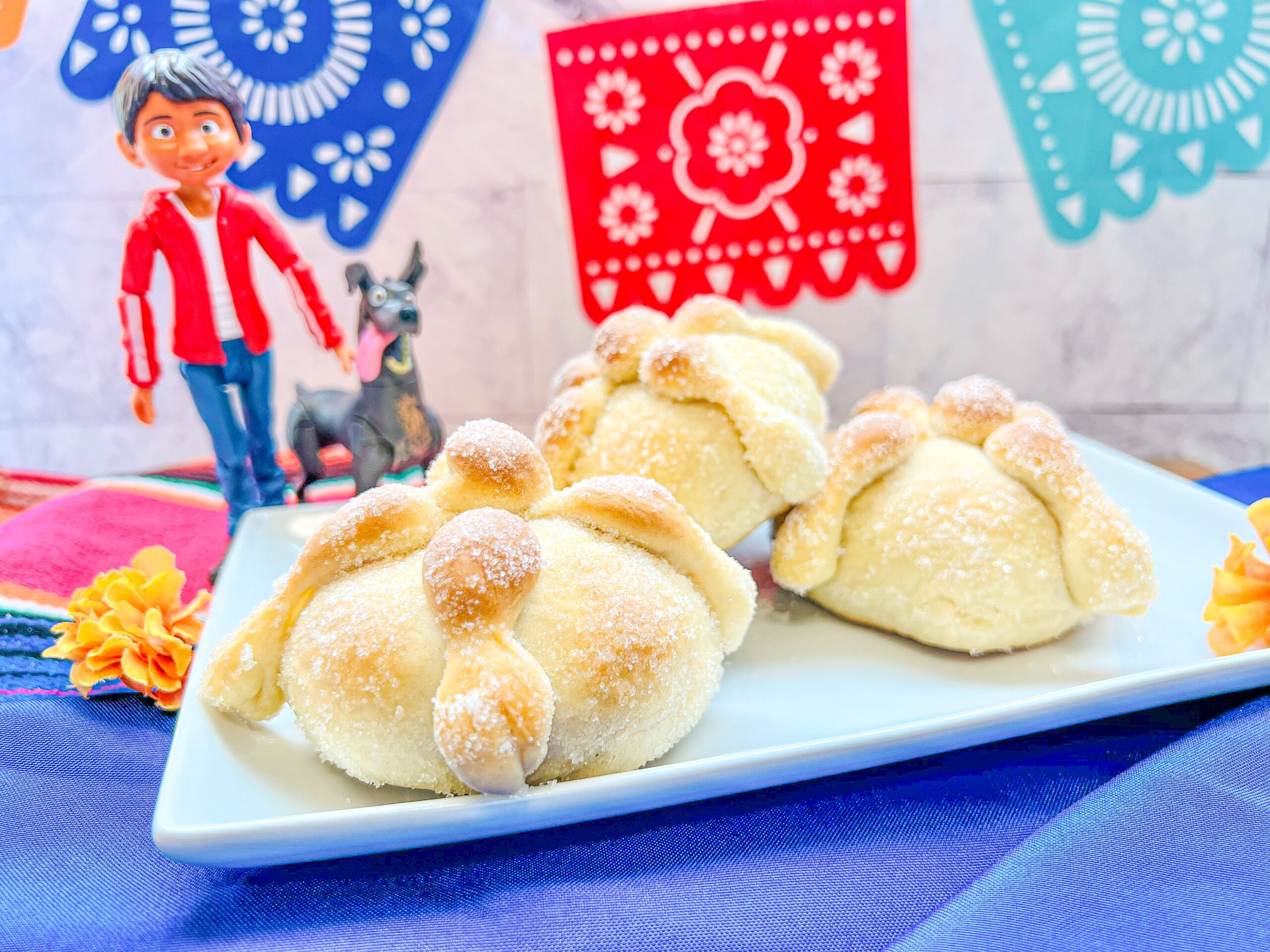 La imagen muestra un plato con mini pan de muerto sobre un camino de mesa con franjas de colores rosa mexicano, blanco, negro y azul rey.