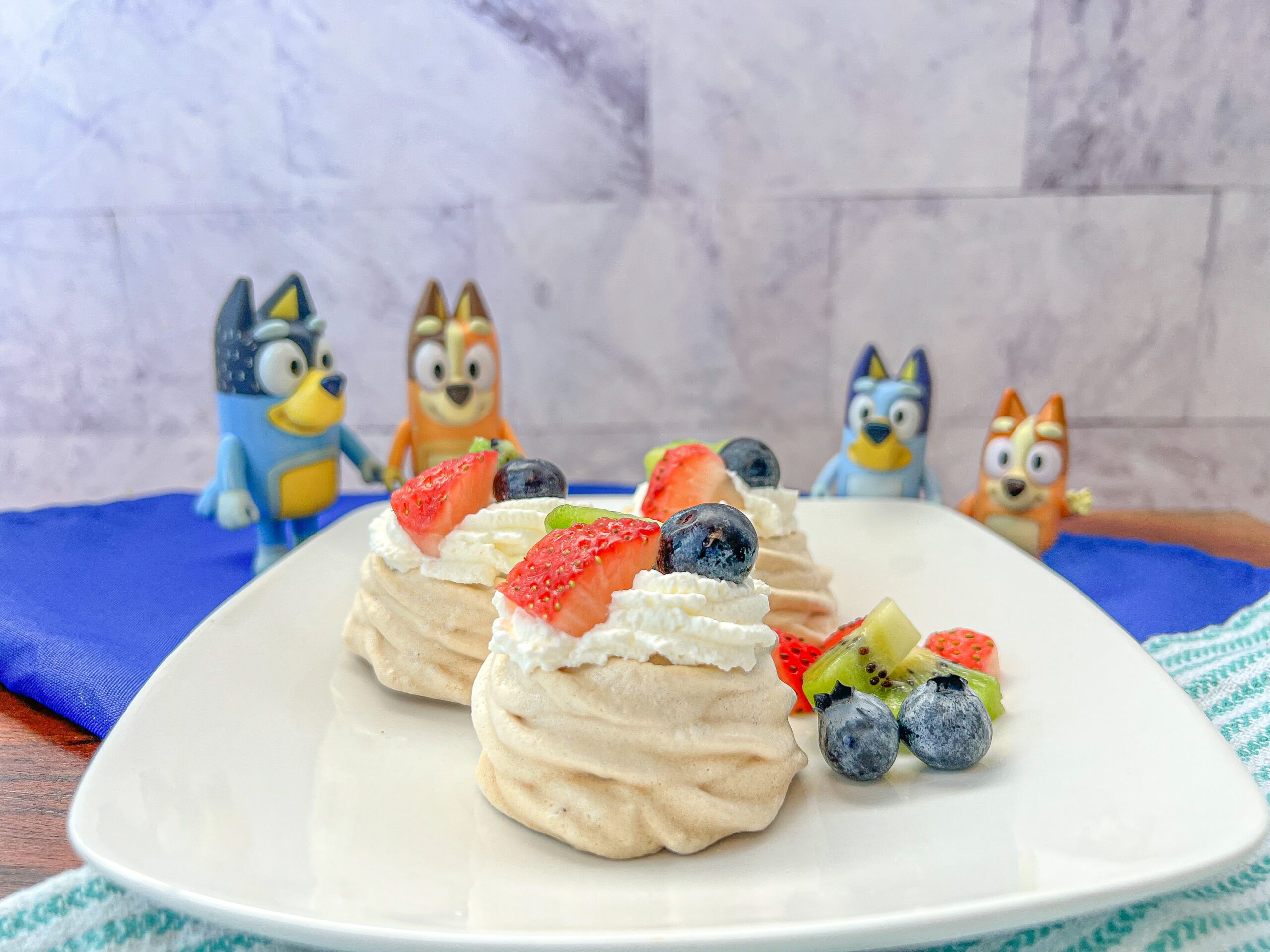 La imagen muestra un plato con tres mini pavlovas decoradas con crema batida, fresas, kiwis y blue berries.