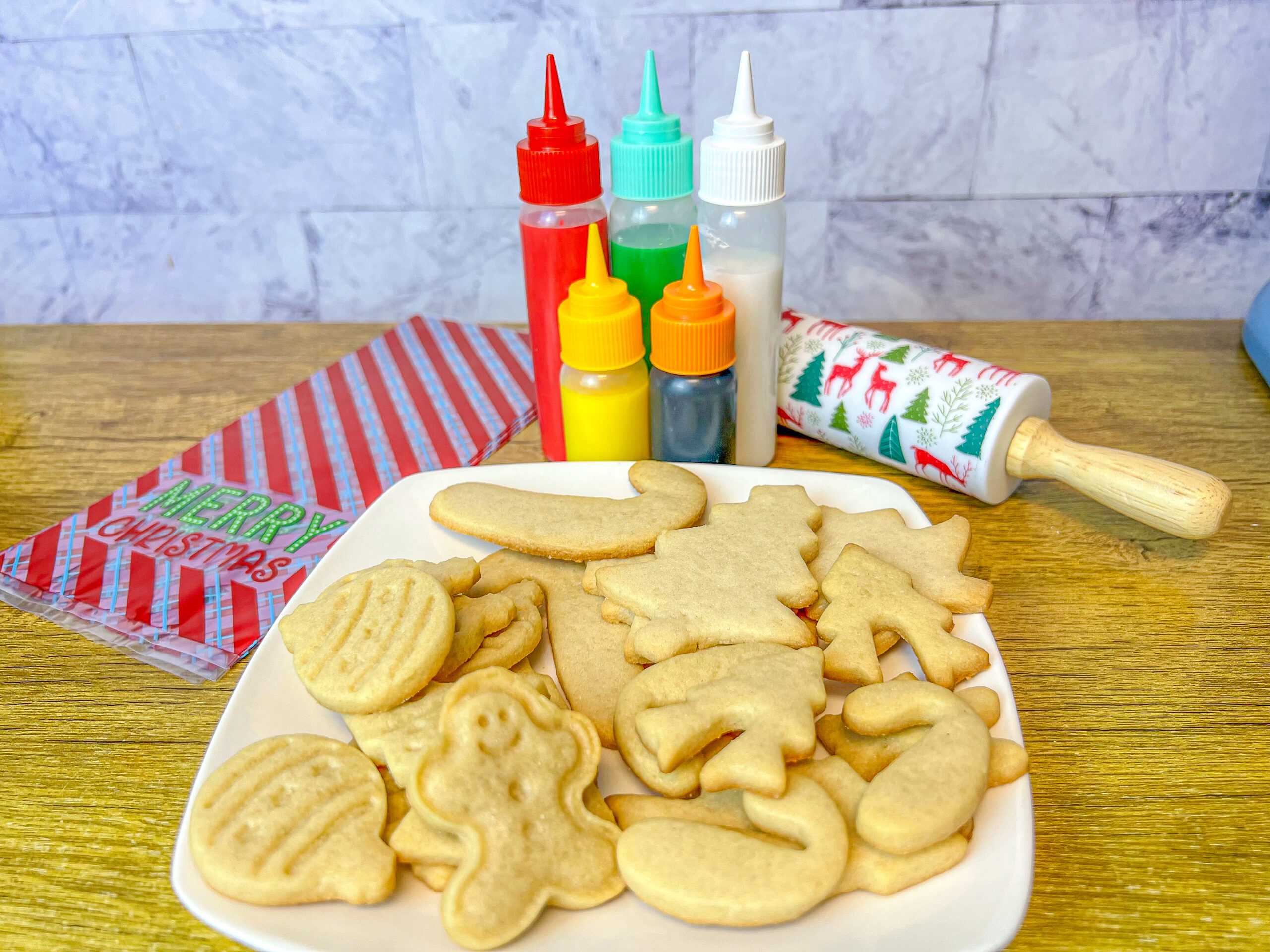 La imagen muestra glaseado para galletas rojo, verde, blanco, amarillo y negro en botellas con aplicador, atrás de un plato con galletas sin decorar.