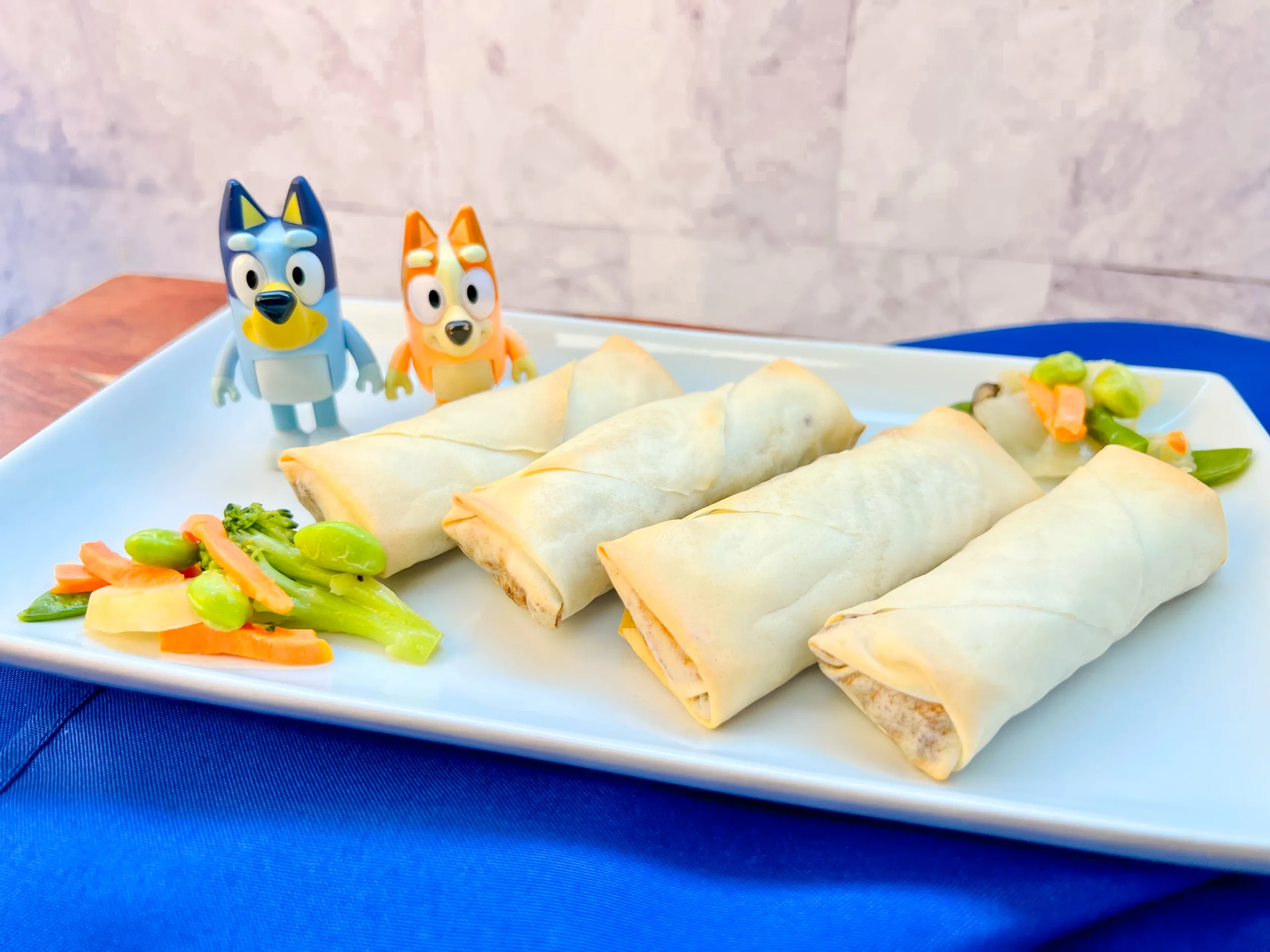 La imagen muestra un plato rectangular con cuatro rollitos primavera acompañado con vegetales y unas figuras de Bingo y Bluey.