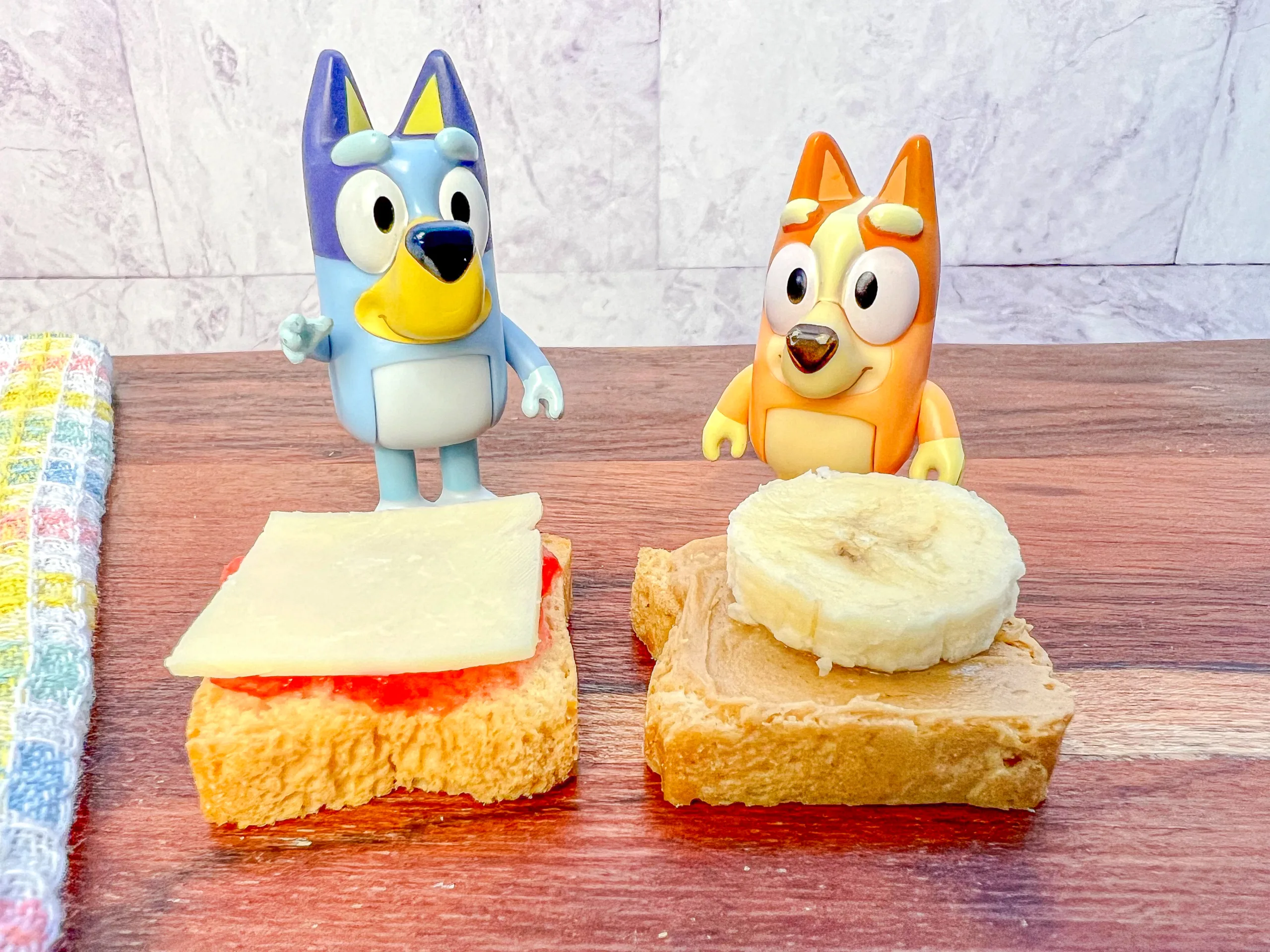 La imagen muestra unos mini panes tostados, uno con mermelada de fresa y queso, y otro con crema de cacahuate y plátano. 