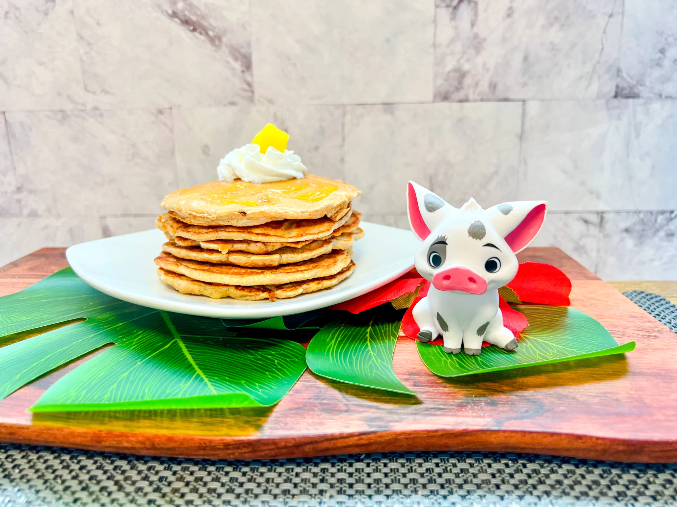 La imagen muestra un plato con pancakes de piña-coco decorados con crema batida y piña inspirados en Moana. Junto a una figura de Púa.