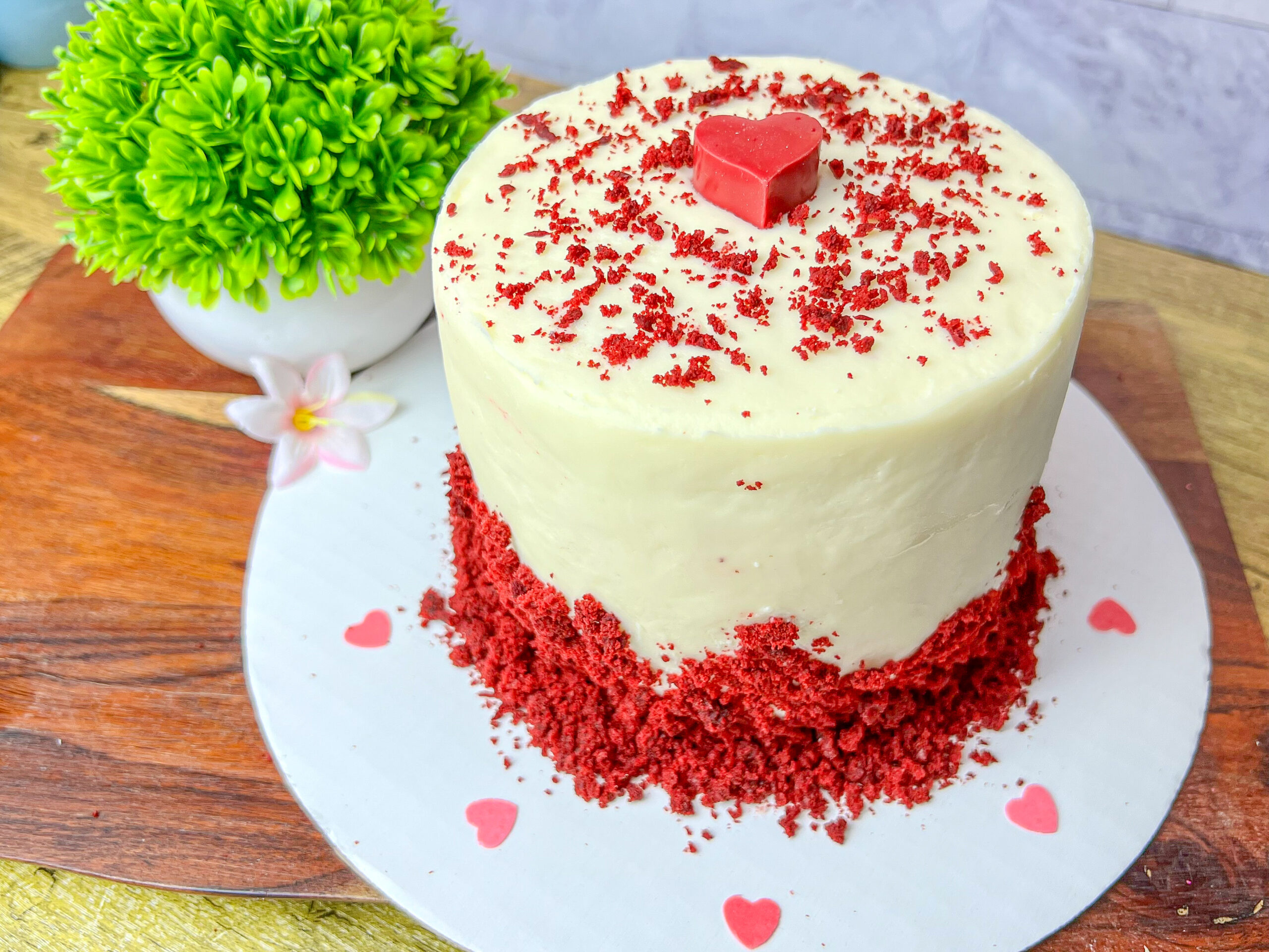 La imagen muestra un mini pastel para San Valentín, hecho de red velvet y cubierto con betún de queso crema, la base tiene morons de pastel cómo decoración y en la parte superior un corazón rojo hecho con caramelo derretido.