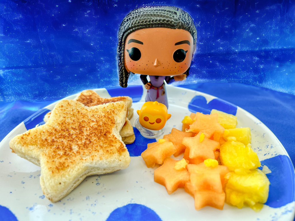 La imagen muestra un plato con dos sándwiches de queso fundido en forma de estrella, acompañados con melón en forma de estrellas y piña. En el plato también se pueden ver 2 figuras de funko de Asha y Estrella como decoración y un fondo azul.