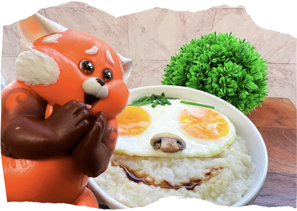 La imagen muestra un plato de congee con carita feliz inspirado en turning red, los ojos están formados por dos huevos estrellados, la nariz con un champiñón salteado, las cejas y el cabello con cebollín y la boca con salsa de soya. Al frente una figura de Mei Mei como Panda Rojo.