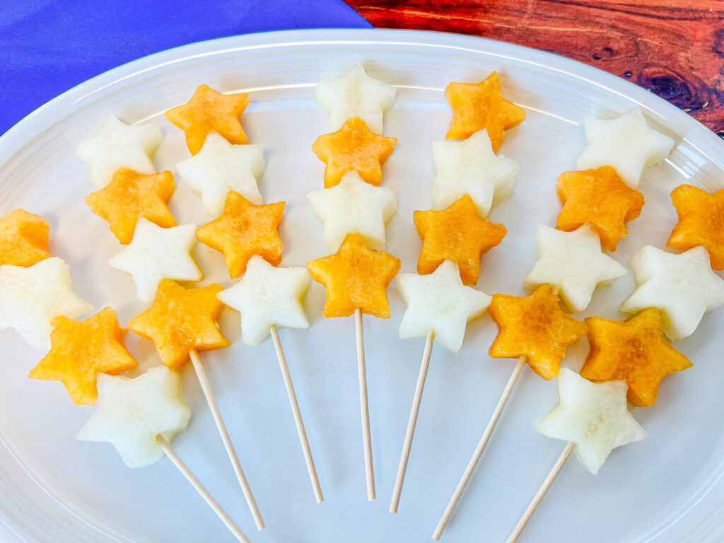 La imagen muestra un plato con brochetas de melón de dos colores en forma de estrellas