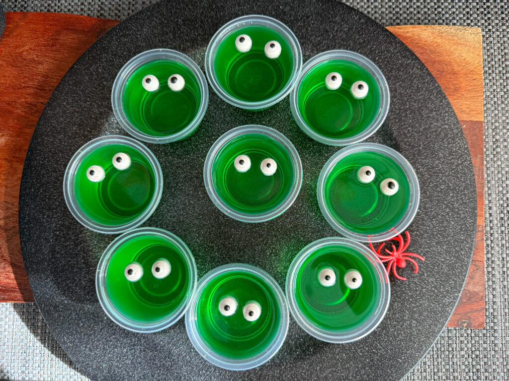La imagen muestra un plato negro con unos vasos de gelatina que simulan monstruos, hechos con gelatina verde con ojos de azúcar.