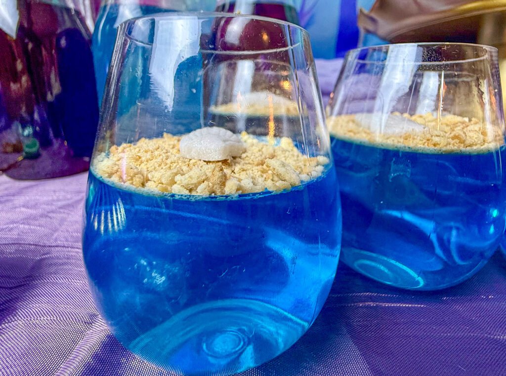 La imagen muestra un par de vasos transparentes vistos de frente con gelatinas marinas, que consisten en gelatina azul con una capa de galletas trituradas encima simulando arena y con un caracol de fondant blanco sobre las galletas.