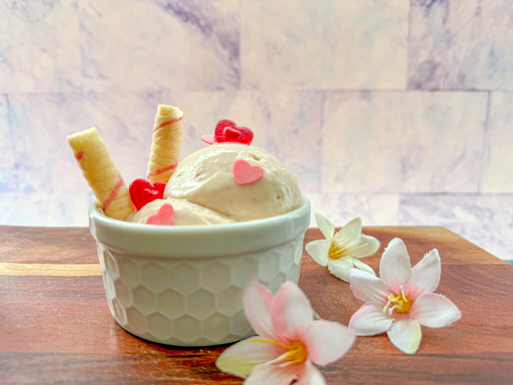 La imagen muestra un helado un helado de fresa casero decorado con dos galletas y corazones rojos y rosas, servido en un ramekin blanco sobre una tabla de madera, con tres flores a lado y un fondo blanco.