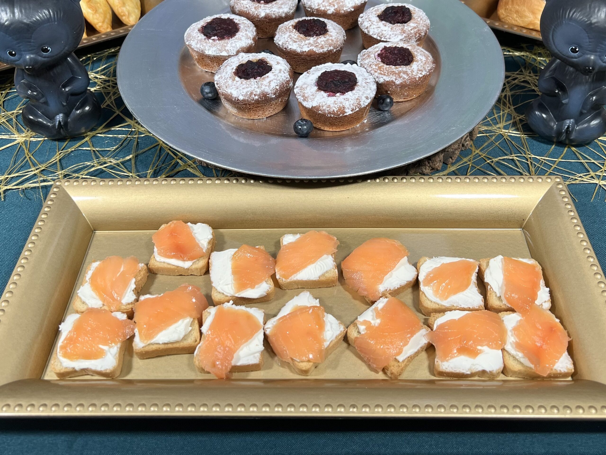 La imagen muestra una charola dorada con canapés hechos con mini panes tostados, una capa de queso crema y una rebanada de salmón ahumado. Sobre una mesa con mantel azul verde y un camino de mesa dorado.